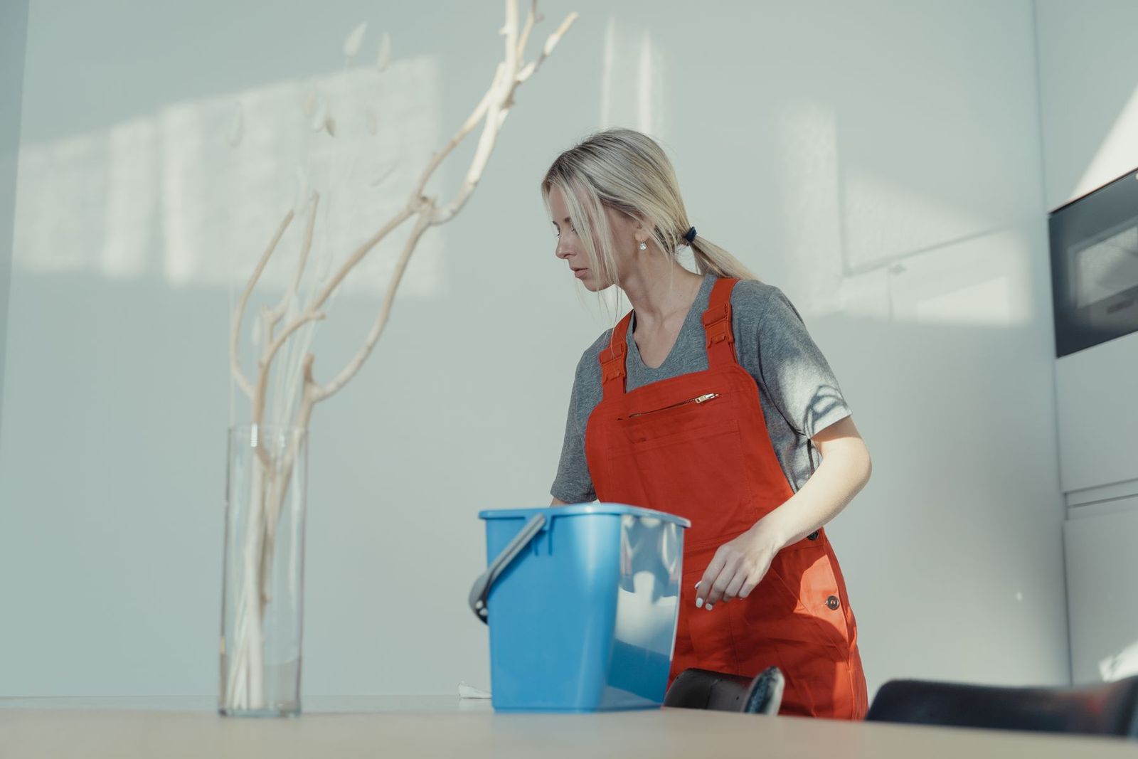 Reinigungskraft in roter Arbeitskleidung mit blauem Eimer in einer Wohnung